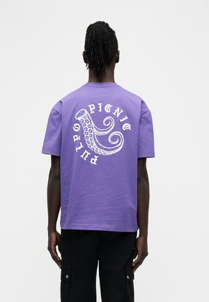 Mann trägt ein violettes T-Shirt mit einem weißen Oktopus-Tentakeldekor und den Worten "PUBLIC PICNIC" auf der Rückseite, kombiniert mit schwarzen Hosen.