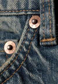 Ύφασμα τζιν με μπλε απόχρωση, διακοσμημένο με χάλκινους πριτσινίσματα και ραμμένες ραφές. Κοντινή λήψη που δείχνει την υφή και τις λεπτομέρειες της υφής.