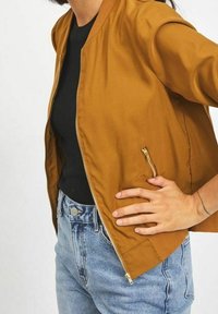 Bomberová bunda v hořčičně žluté látce se zipem v přední části, žebrovaným límcem a bočními kapsami; kombinována s modrými džínami s vysokým pasem.