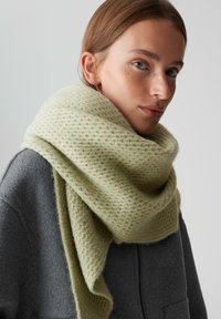 Jeune femme portant un grand foulard texturé de couleur vert clair enroulé autour de son cou et de ses épaules, sous un manteau gris à fermeture éclair.