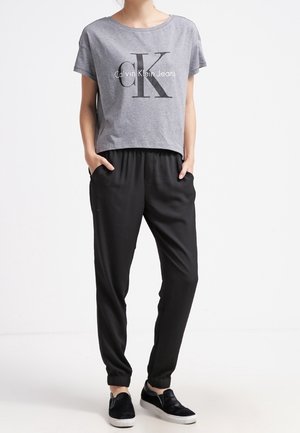 Osoba ubrana w szarą krótką koszulkę Calvin Klein Jeans, czarne spodnie dresowe z elastyczną talią oraz czarne wsuwane trampki z białą podeszwą.