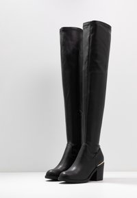 Botas altas de cuero negro con tacón, con cremallera lateral, puntera redondeada y tacón cuadrado, posicionadas de pie sobre una superficie blanca contra un fondo liso.