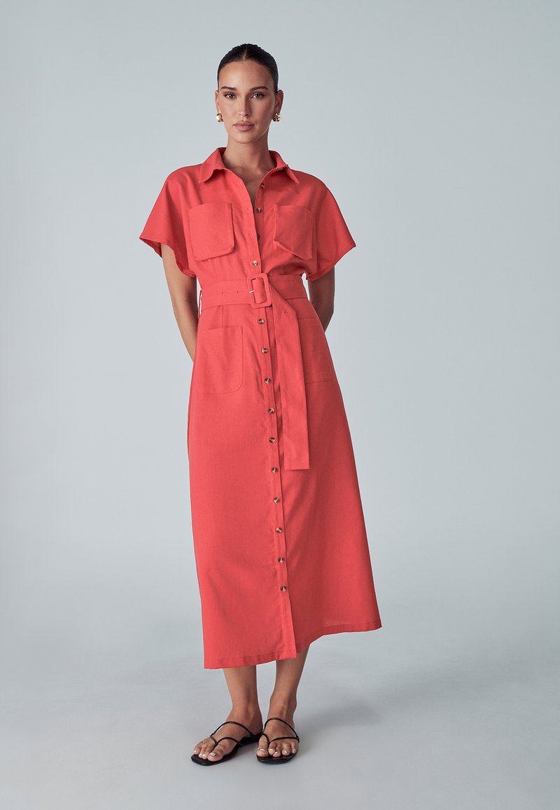 Koronkowa sukienka koszulowa w kolorze koralowym z krótkimi rękawami, z przodu zapinana na guziki, z dwiema kieszeniami na piersi oraz paskiem w talii. Materiał wygląda na lekki o gładkiej fakturze.