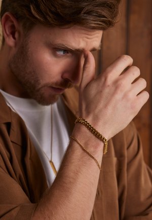 Gold-tone chain bracelet with a textured link design on a wrist, layered with a thinner gold-tone chain, set against a wooden background.