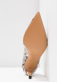 Chaussure à talon haut à bout pointu avec semelle tan et motif en peau de serpent noir et blanc appuyée contre un mur blanc.
