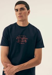 Granatowa koszulka z krótkim rękawem, wykonana z bawełny, z czerwoną grafiką na przodzie, na której widnieje napis "HENDERSON Malibu Beach CALI.".