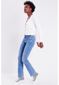Gilet texturé blanc, jean flare taille haute bleu clair avec trois accents de boutons métalliques, et baskets à carreaux noir et blanc.