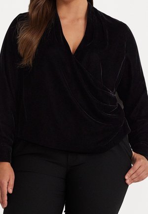 Femme portant un haut cache-cœur en velours noir à manches longues et un pantalon noir, main posée sur la hanche, sur fond blanc.