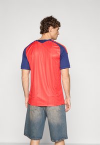 Röd och blå sportt-shirt med korta ärmar, med en texturerad rygg med vertikal sömnad och kontrasterande axeldetaljer.