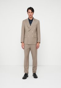 Μπεζ διπλής όψης κοστούμι με ασημένια κουμπιά, στενές παντελόνες και σκούρο πουκάμισο από κάτω, ολοκληρωμένο με μαύρα δερμάτινα παπούτσια.