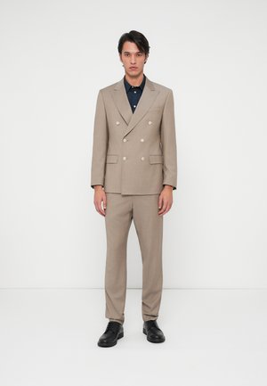 Beige kaksirivinen puku, jossa on hopeiset napit, slim fit -housut ja tummanvärinen paita alla, viimeistelty mustilla nahkakengillä.