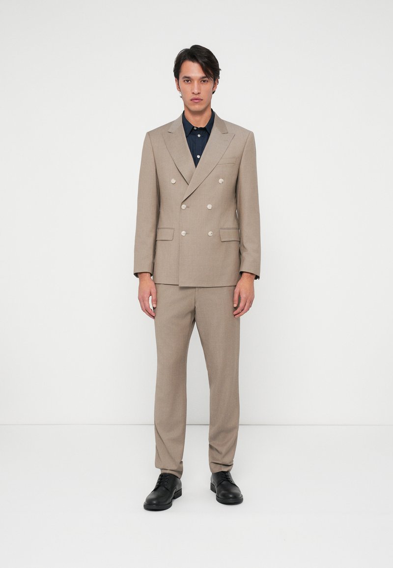 Μπεζ διπλής όψης κοστούμι με ασημένια κουμπιά, στενές παντελόνες και σκούρο πουκάμισο από κάτω, ολοκληρωμένο με μαύρα δερμάτινα παπούτσια.