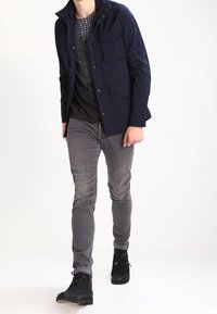 Mörkblå jacka med flera framfickor, bärs över en svart t-shirt med nätmönster, kombinerad med grå slim-fit jeans och svarta hög-top sneakers.