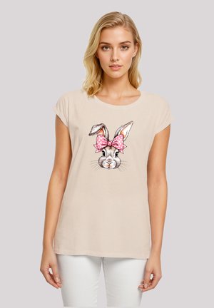 Beige short-sleeve t-shirt featuring a rabbit graphic with large ears and a pink polka dot bow. Soft fabric with a relaxed fit.