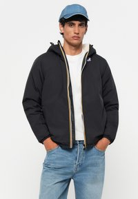 Veste isolée noire avec une capuche, zip jaune et patch de logo. Associée à une chemise blanche et un jean bleu clair. Casquette en denim portée.