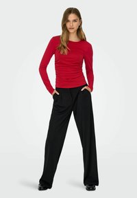 Rode top met lange mouwen en geplooide textuur, gecombineerd met losse zwarte broek. Model staat met handen in de zakken en draagt zwarte schoenen.