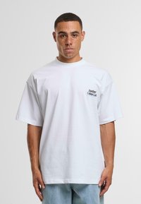 Camiseta de algodón blanca de gran tamaño con cuello redondo y mangas cortas, que presenta un pequeño logotipo negro bordado en el lado izquierdo del pecho.