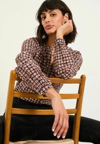 Flower-patterned blouse in soft fabric, featuring button-down front, long puffed sleeves, and gathered details. Model seated on a wooden chair.