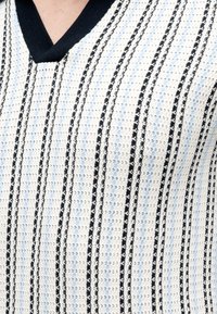 Gros plan d’une personne portant un haut en maille texturée avec des rayures verticales noires, blanches et bleu clair et un col en V noir.