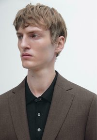 Giacca marrone su misura sopra una camicia nera con colletto e tre bottoni, caratterizzata da una texture liscia e linee pulite.