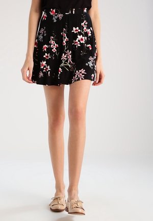 Vrouw die zwarte korte broek met bloemenprint en plooien draagt, gecombineerd met beige loafers met kwastjes, staand tegen een effen lichte achtergrond.