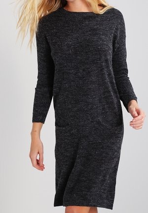 Kobieta ubrana w ciemnoszarą, dzianinową sukienkę z długim rękawem, sięgającą do kolan, z okrągłym dekoltem i subtelną fakturą.