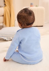 Světlomodrý pletený dvoudílný komplet s žebrovanými manžetami a límcem, doplněný jemnými bílými detaily, na sobě má sedící kojence s kudrnatými vlasy.