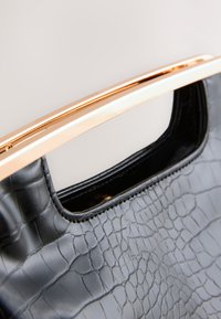 Melnas ādas rokas soma ar krokodila tekstūru, ar spīdīgu zeltīti metāla rokturi. Interjers redzams caur atvērtu augšu.