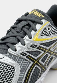 Gray athletic shoe with mesh and synthetic materials, highlighted with yellow accents, featuring a textured surface and intricate lacing.