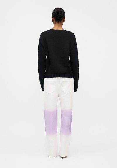 Γυναίκα που στέκεται με την πλάτη προς το μέρος μας, φοράει μαύρο μακρυμάνικο πουλόβερ και λευκό παντελόνι φαρδύ στο πόδι με μοβ διαβάθμιση από το μέσο του μηρού έως τους αστραγάλους.