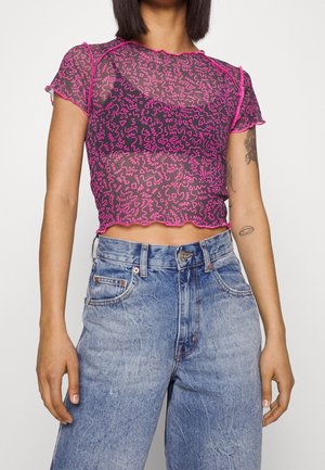 Läpinäkyvä musta toppi, jossa on pinkki abstrakti kuvio, aaltoilevat reunat ja lyhyet hihat, yhdistettynä korkeavyötäröisiin farkkuihin.