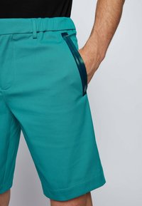 Tyrkysové šortky s elastickým pasem, které mají elegantní boční kapsu s kontrastním tmavě modrým akcentem a hladkým, lehkým materiálem.