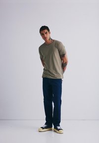 Olivengrønn T-skjorte med korte ermer, blå denimjeans og svarte sneakers med hvite snørebånd. Modellen står mot en hvit bakgrunn.