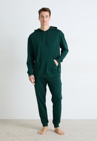 Hanorac lung, cu maneci lungi, de culoare verde închis, cu un buzunar frontal, asociat cu pantaloni taperati asortați. Fabrică moale, elastică, detalii minime, croială lejeră.