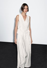 Kobieta stojąca w pobliżu białej ściany, ubrana w bezrękawnikowy biały top z owijanym przodem oraz szerokie białe spodnie, z krótkimi ciemnymi włosami i neutralnym wyrazem twarzy.