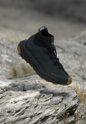 Schwarzer wasserdichter Wanderschuh mit robuster brauner Sohle, mittelhohem Design und Schnellverschluss-System, der über felsigem Gelände im Freien schwebt.