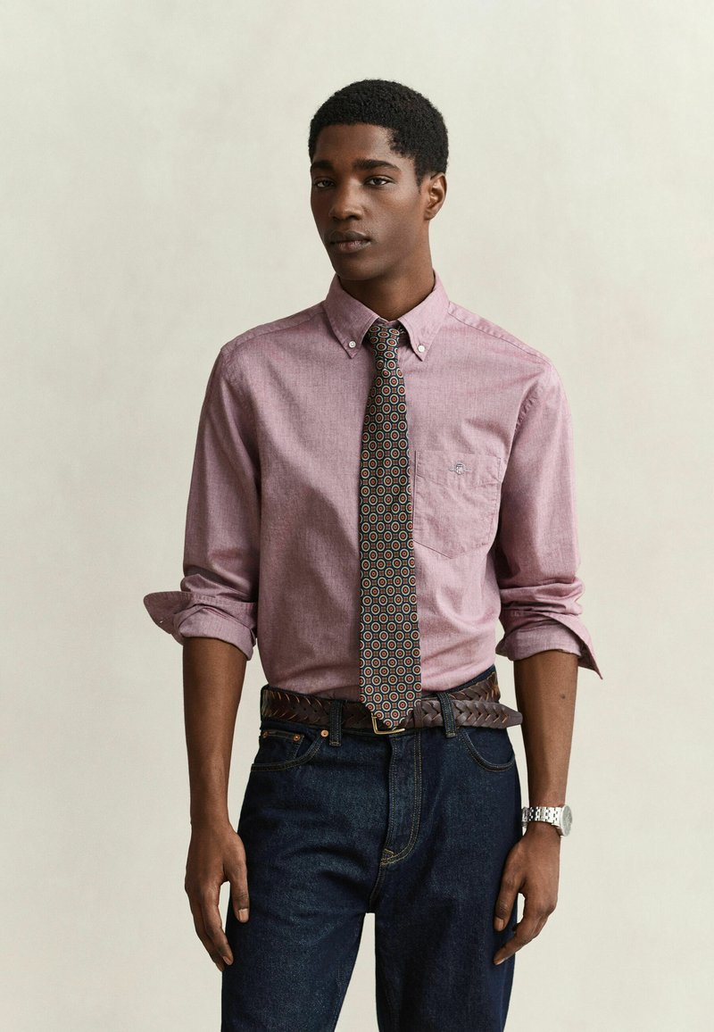 Jeune homme portant une chemise rose boutonnée aux manches retroussées, une cravate à motifs, un jean foncé, une ceinture marron tressée et une montre-bracelet argentée, debout devant un fond uni.