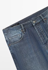 Modré džínové kalhoty s přední kapsou, poutky na opasek, kovovým knoflíkem a viditelným vnitřním páskem s logem na bílém pozadí.