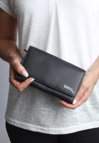 Black leather wallet with a smooth texture, featuring a fold-over design and subtle logo. Held in a person's hand, against a light background.