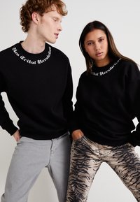 Sudaderas negras con el texto blanco "Revolución Neo Tribal" en el escote, combinadas con pantalones gris y con estampados. Acabado de tela texturizada.