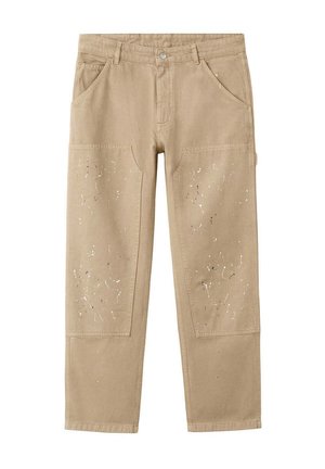 Osoba nosząca beżowe robocze spodnie poplamione farbą z bocznymi kieszeniami oraz ciemnoniebieską bluzkę z długim rękawem, jedną ręką w kieszeni, drugą luźno opartą.