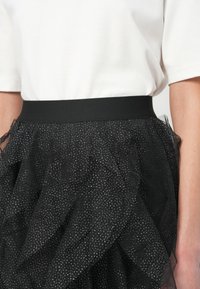 Juoda tiulio sijonas turi plačią liemenėlę, sluoksniuotas volanas ir subtilų blizgantį raštą visame sijone. Audinys yra lengvas ir tekstūrinis.