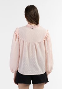 Lichtroze blouse met gestructureerde stippen, gerimpelde kraag en manchetten, lange mouwen en een geplooide achterzoom, gedragen met donkere shorts.