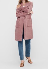 Langer, rosa Cordcardigan aus Cord mit zwei Vordertaschen, offenem Frontdesign und gerippter Struktur. Kombiniert mit blauer Jeans und braunen Sandalen.