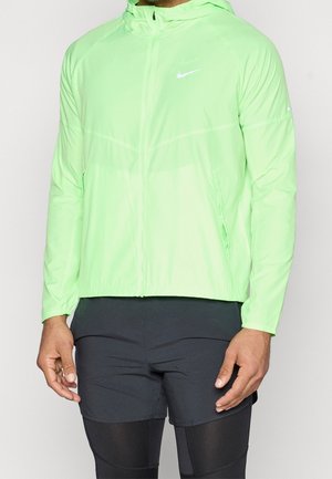Homme portant une veste à capuche zippée Nike vert clair, un short noir et des leggings noirs, debout devant un fond uni.