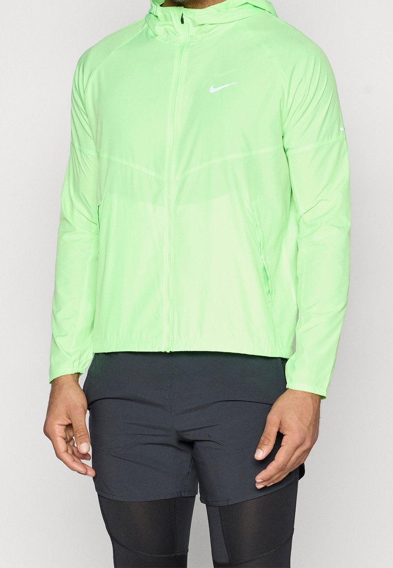 Homme portant une veste à capuche zippée Nike vert clair, un short noir et des leggings noirs, debout devant un fond uni.