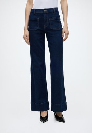 Vue de face d'une personne portant un jean bleu foncé à jambes larges avec de grandes poches avant et des chaussures noires à talons hauts.