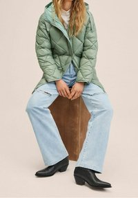 Chaqueta acolchada verde con capucha, combinada con vaqueros azul claro con un detalle rasgado y botines negros puntiagudos.