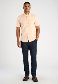 Korte mouwen, perzikkleurig knoopshirt met een gestructureerde afwerking, gecombineerd met marineblauwe broek en bruine schoenen, staand tegen een neutrale achtergrond.