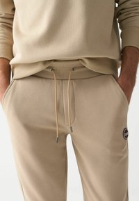 Sudadera beige y pantalones deportivos a juego con cinturilla ajustable, puños acanalados y un parche de logo circular en la pierna del pantalón. Textura de tela suave.